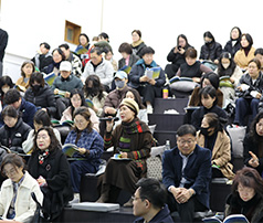 계단식 좌석에 앉은 많은 청중 중 한 여성이 마이크를 들고 질문이나 소감을 발표하고 있으며, 주변 사람들이 경청하는 모습