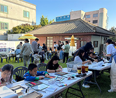 맑은 날 야외 잔디밭 테이블에 앉아 어린이들이 집중해서 그림을 그리거나 만들기를 하고, 뒤편 한옥 건물 앞에서 사람들이 자유롭게 활동하는 모습