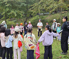 숲속 평지에서 아이들과 선생님들이 둥글게 원을 그리며 손을 잡거나 마주 보고 서서 야외 레크리에이션 활동을 즐기는 모습